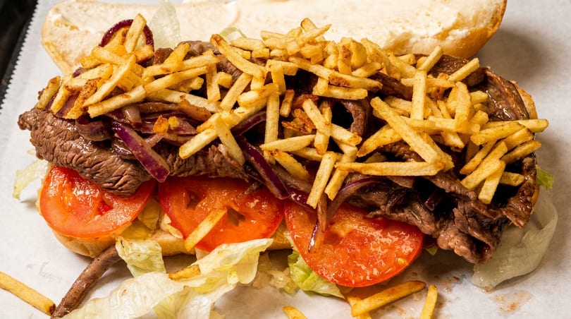 Steak sandwich with french fries, red onions, tomatoes, and lettuce on a white bun.