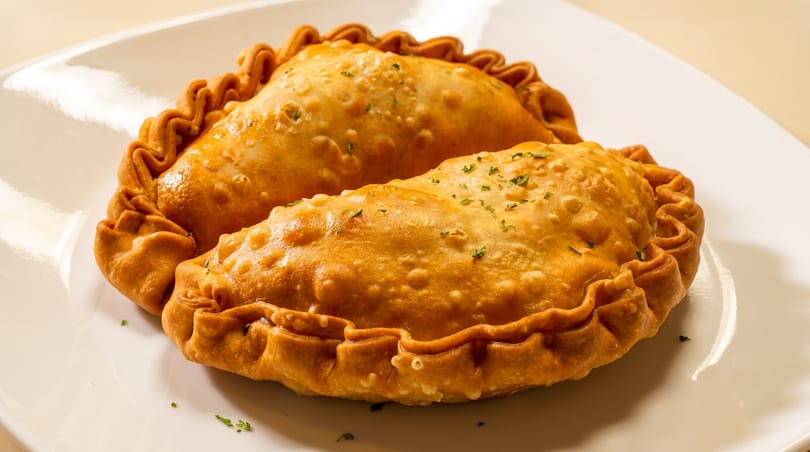 Two golden-brown empanadas with crimped edges and herbs on a white plate.