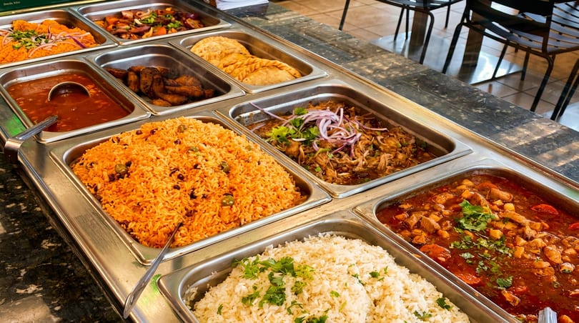 Restaurant buffet with various hot dishes including rice, stews, meats, empanadas, and plantains.