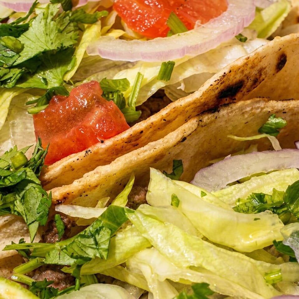 Close-up of three tacos filled with ground meat, lettuce, red onion, cilantro, and tomato, with lime wedges.