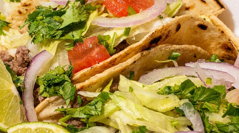 Close-up of three tacos filled with ground meat, lettuce, red onion, cilantro, and tomato, with lime wedges.